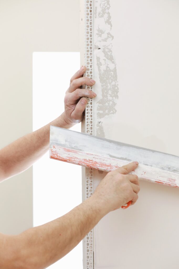 Builder in protective mask using a trowel to add plaster. Plastering wall with putty-knife, close